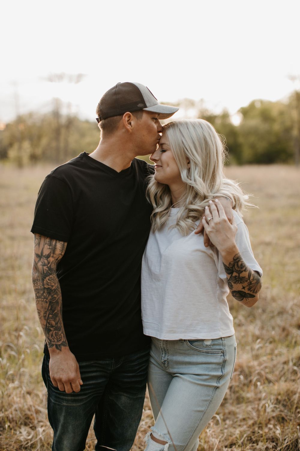 Tattooed couple almost kissing in golden field, man in black shirt and cap leaning toward smiling woman.