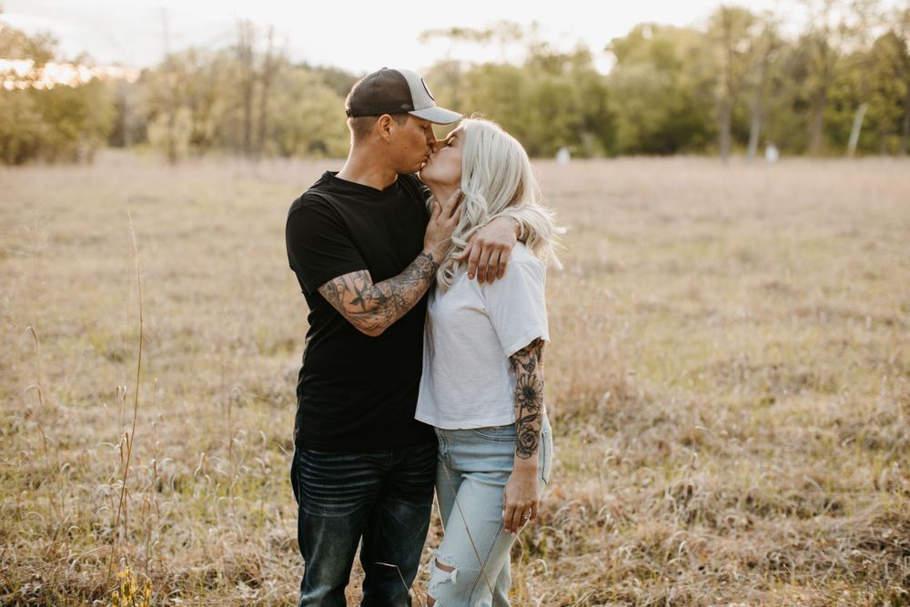 Tattooed couple kissing in open field, man in black shirt embracing woman in white tee, sunny outdoors.