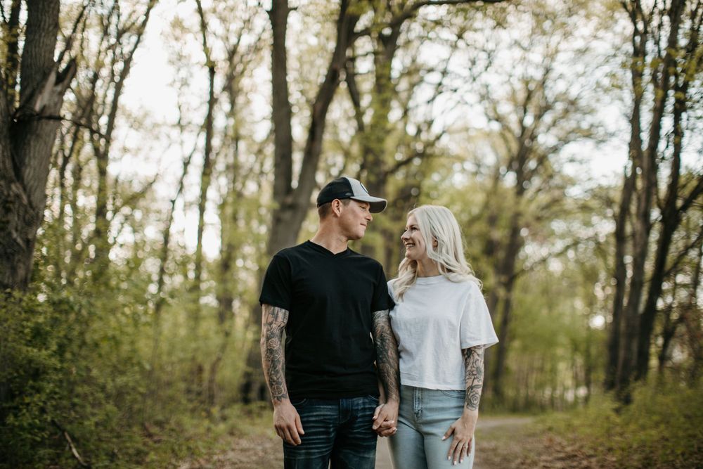 Smiling tattooed couple holding hands on a forest path during a sunny outdoor engagement session.
