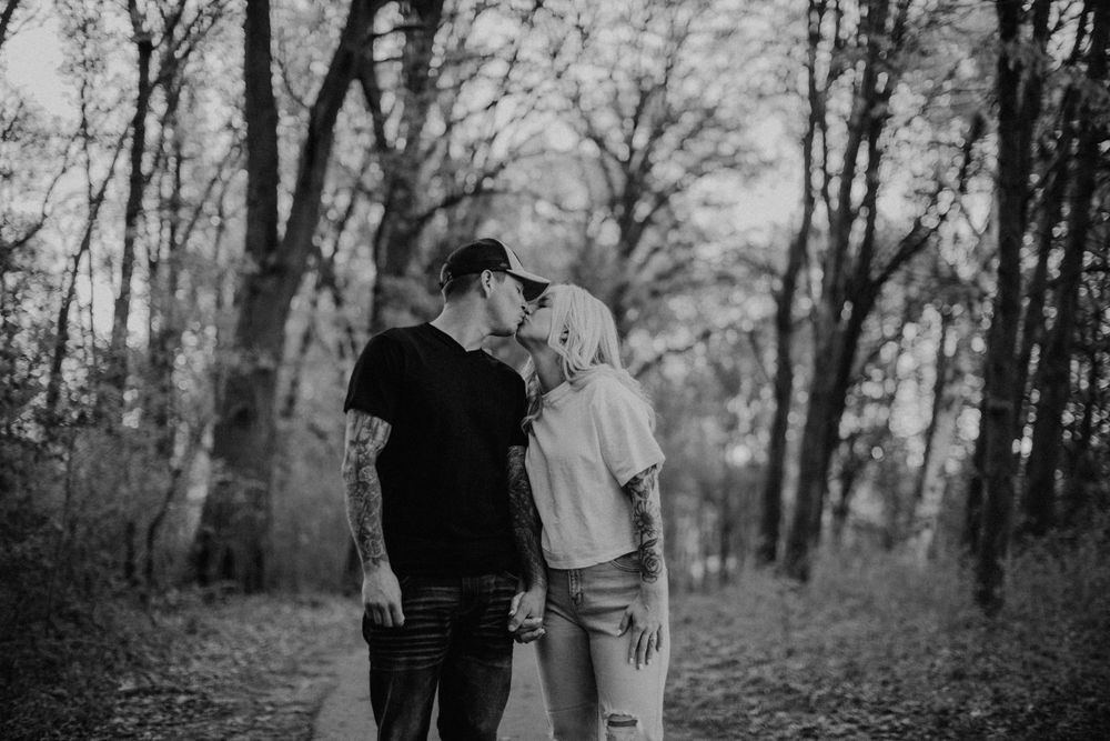 Tattooed couple sharing a tender kiss on a woodland path in black and white.