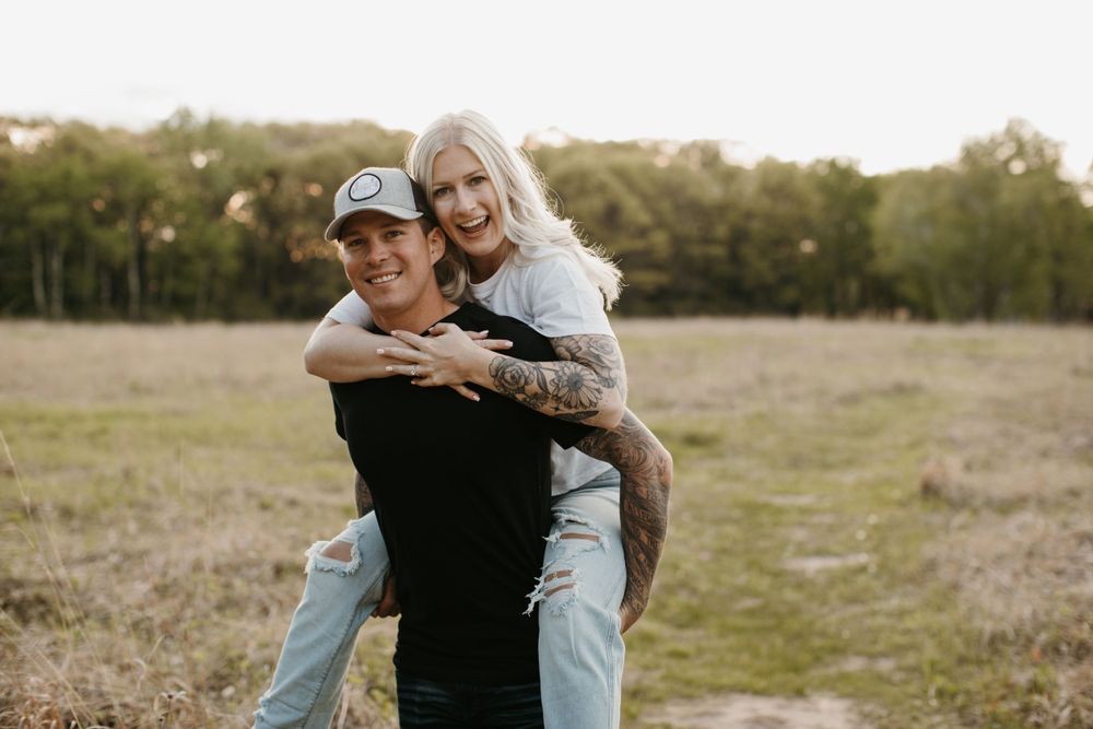 Smiling man giving tattooed blonde woman a piggyback ride in a sunny open field.