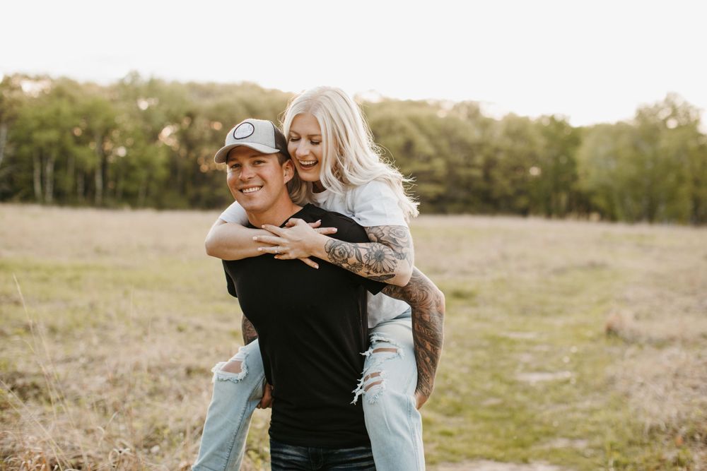 Couple laughing during a piggyback ride in a sunny meadow during outdoor photo session.