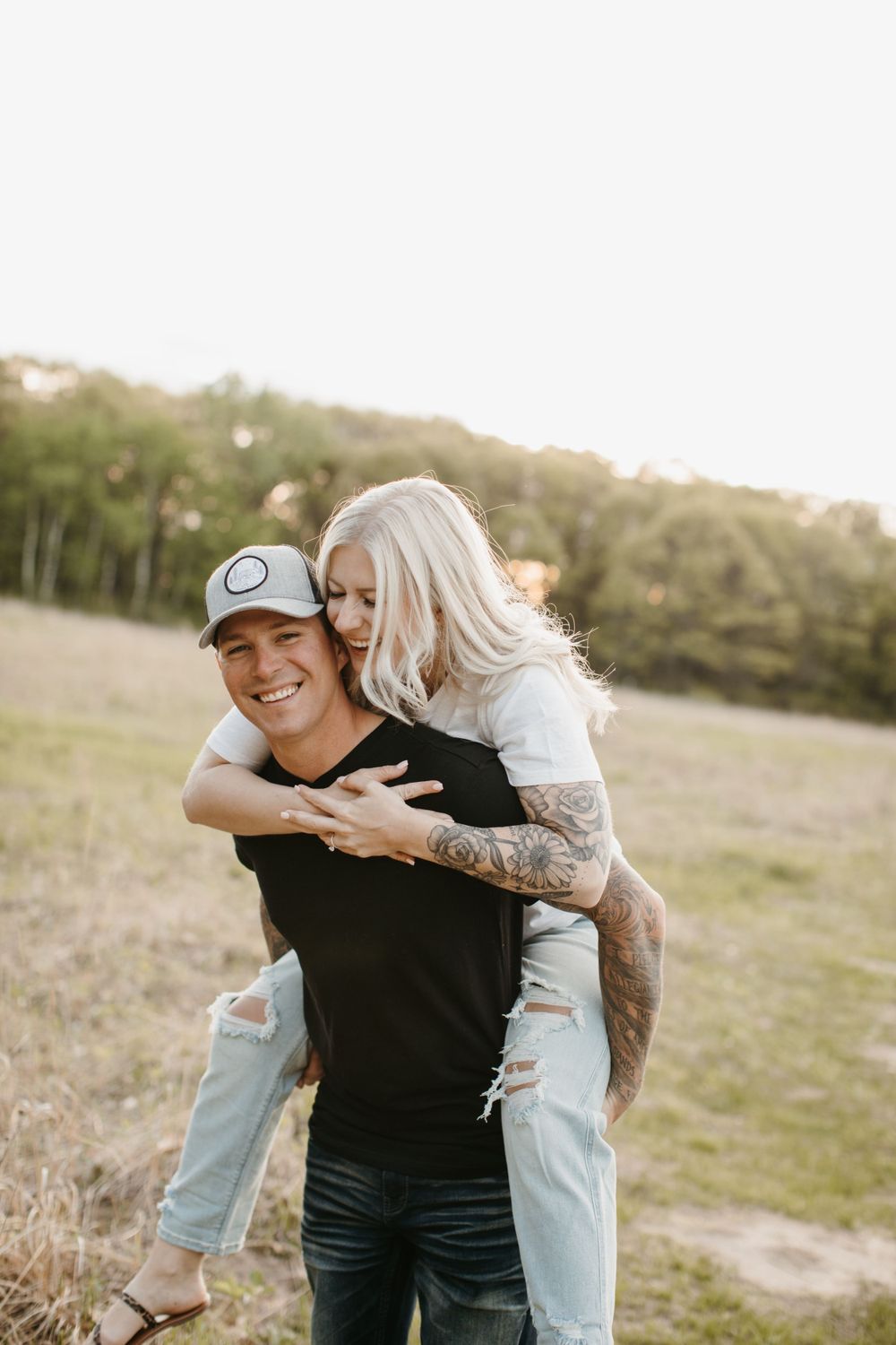 Man giving laughing tattooed blonde woman a piggyback ride in a golden sunlit field.