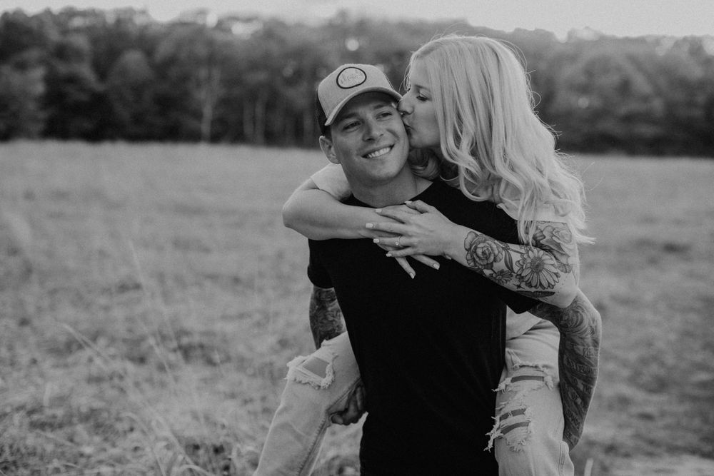 Man giving tattooed blonde woman a piggyback ride, black and white romantic field photo.