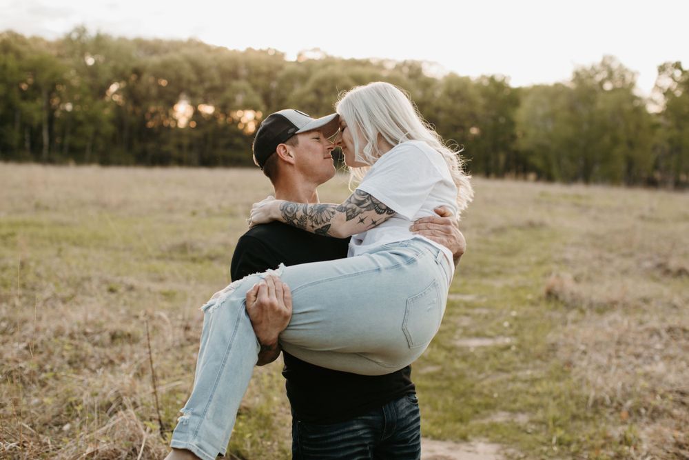 Man lifting and kissing tattooed blonde woman in a romantic outdoor field setting.