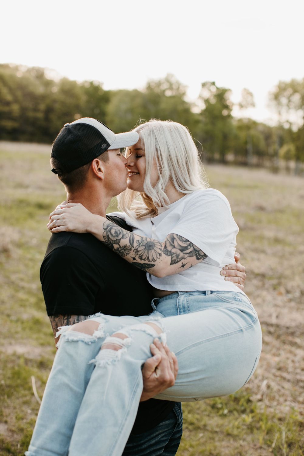 Tattooed couple kissing while man lifts woman in a sunny outdoor meadow setting.