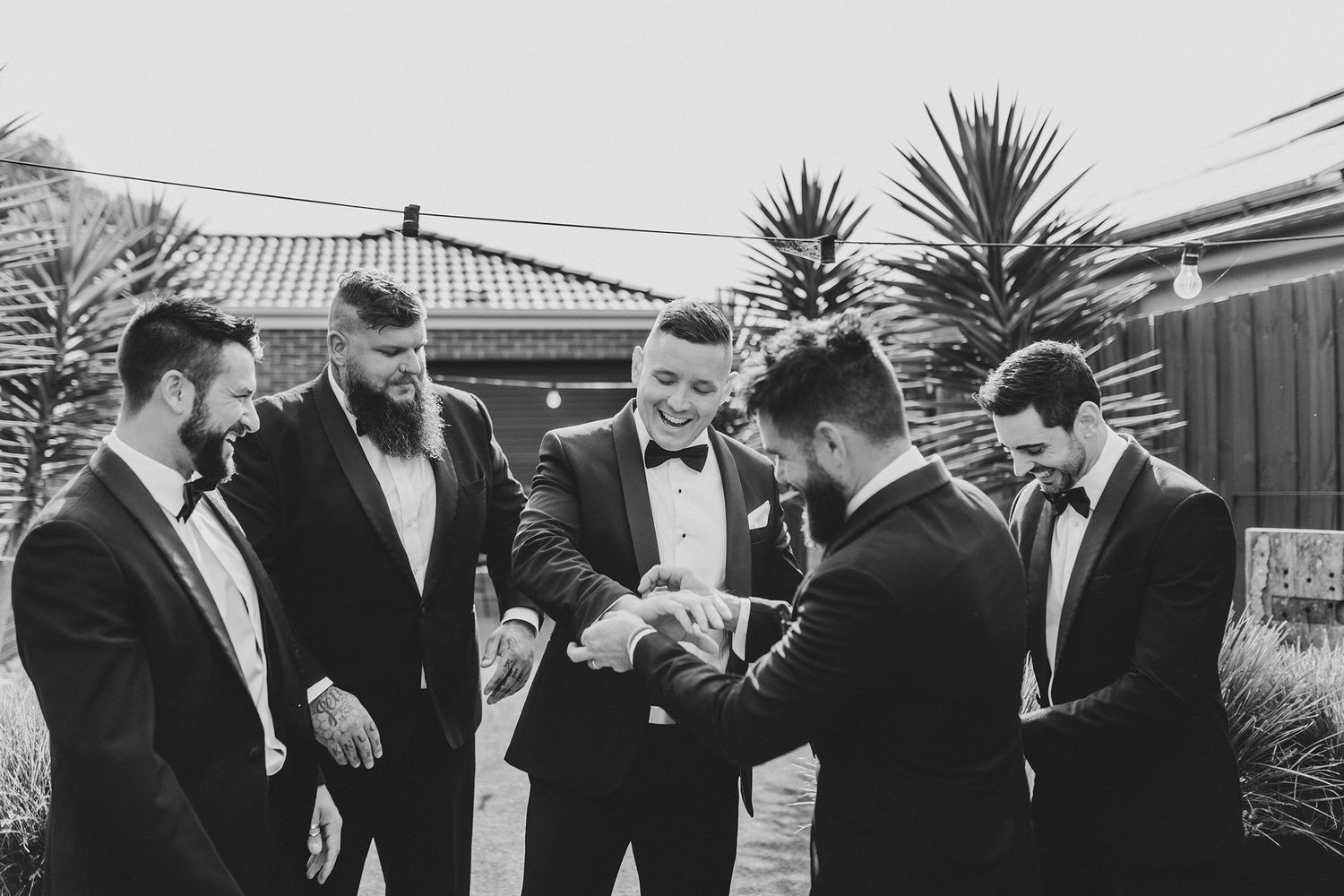 Groomsmen gather in formal black tuxedos examining something together in an outdoor setting.