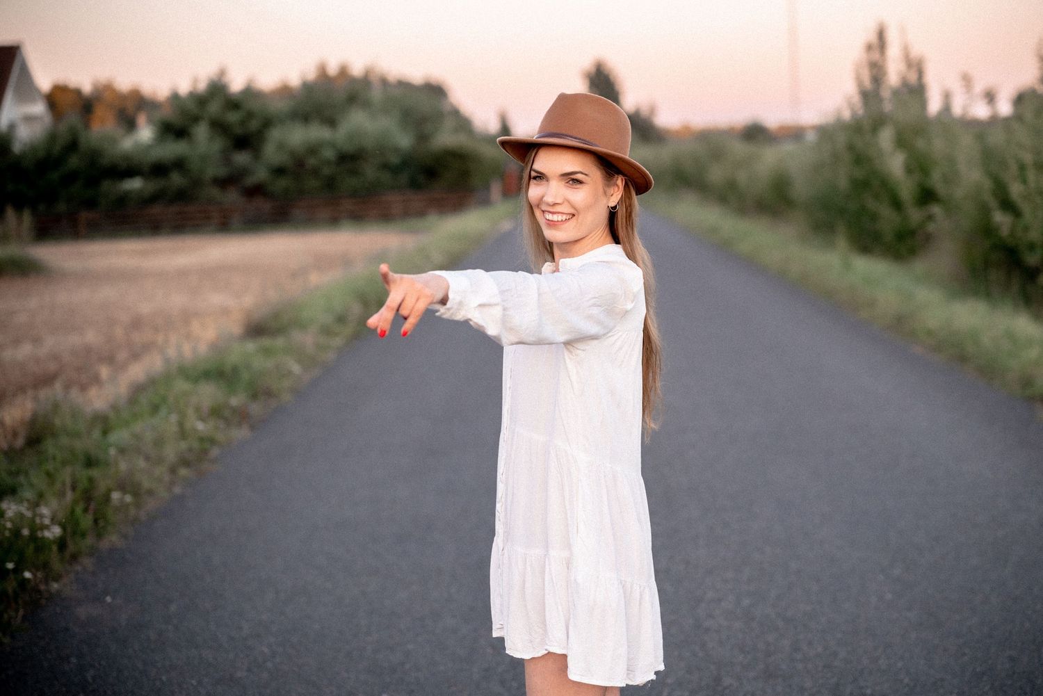 A person in a flowing white dress and brown hat points while standing on a rural road.