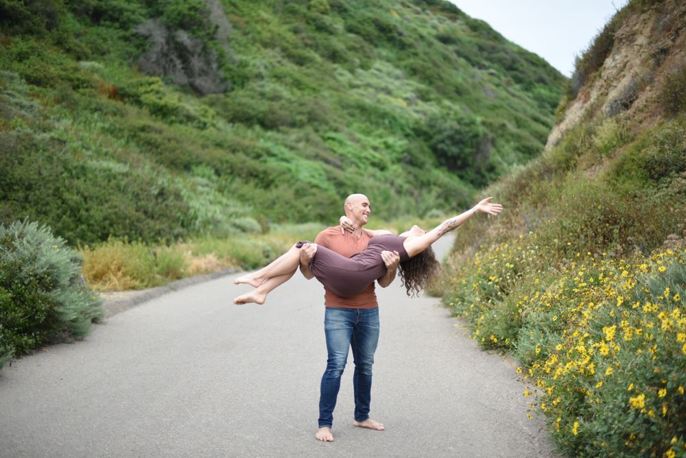 Two people playfully pose on a coastal path with green hills and yellow wildflowers.