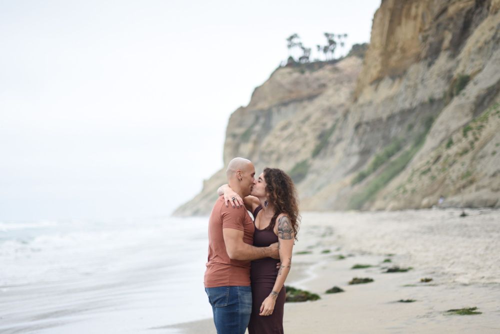 Couple embracing on beach with dramatic coastal cliffs in background.