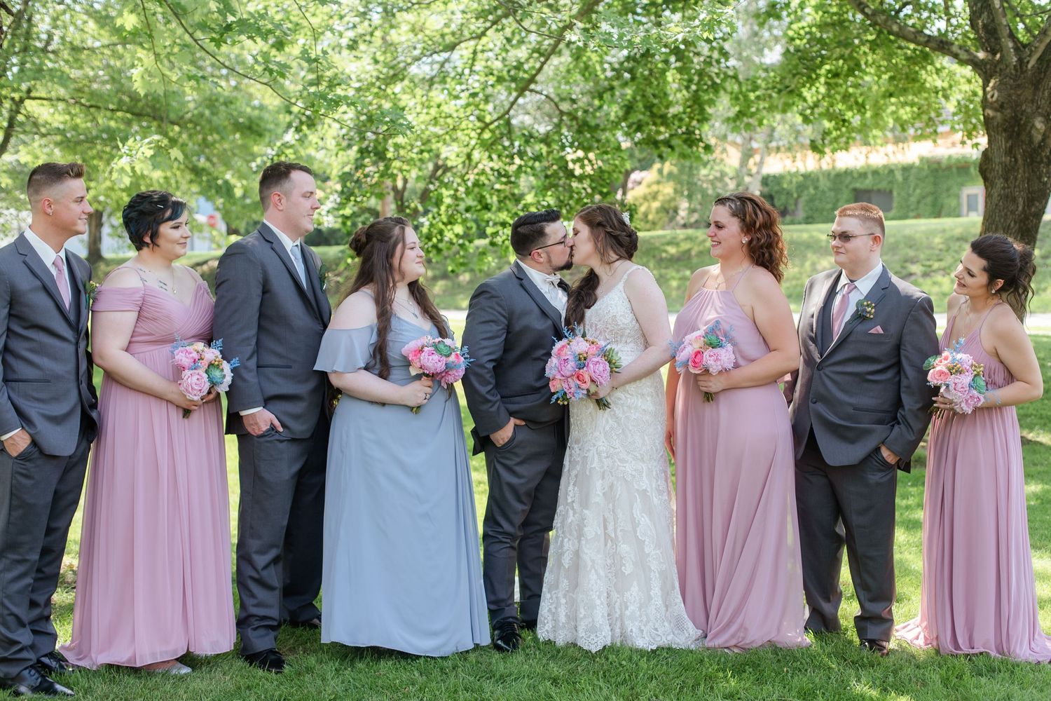 Large wedding party in pink and blue dresses with dark suits poses in garden setting.