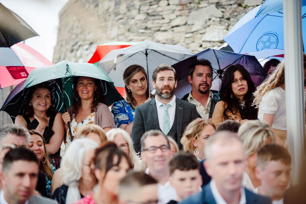 Wedding guests huddle under umbrellas during an outdoor ceremony on a rainy day.