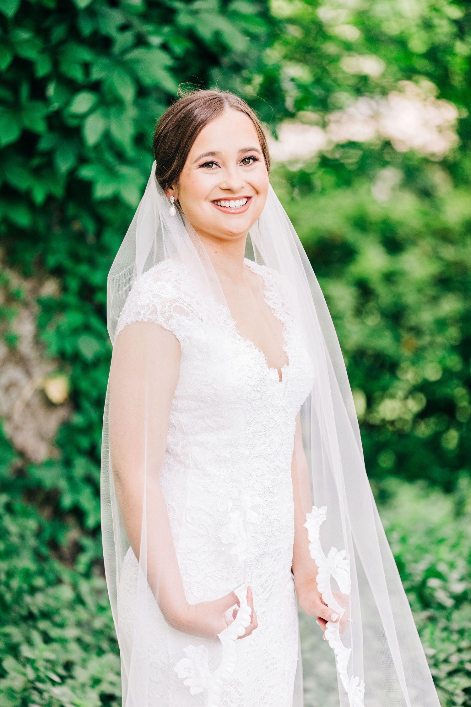 A bride in a white lace wedding dress and veil poses outdoors against lush greenery.