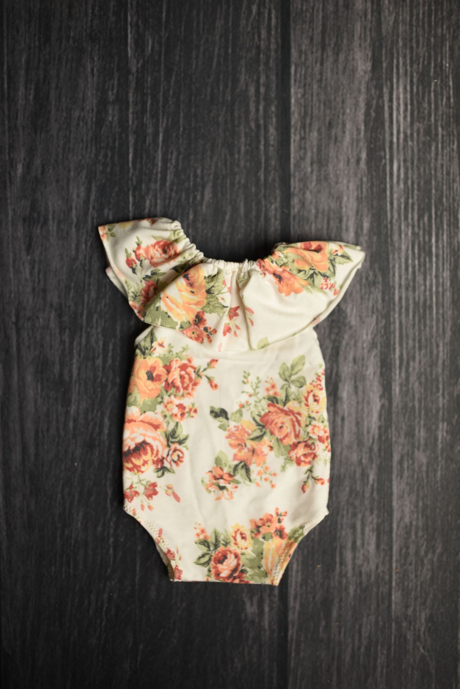 Floral print baby bodysuit with ruffled sleeves on dark wooden background.