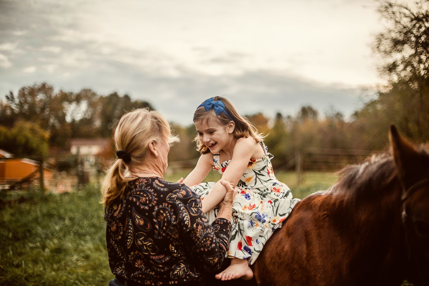 a young girl wearing a floral dress sits sideways on a horse; her grandmother is hugging her as they laugh together