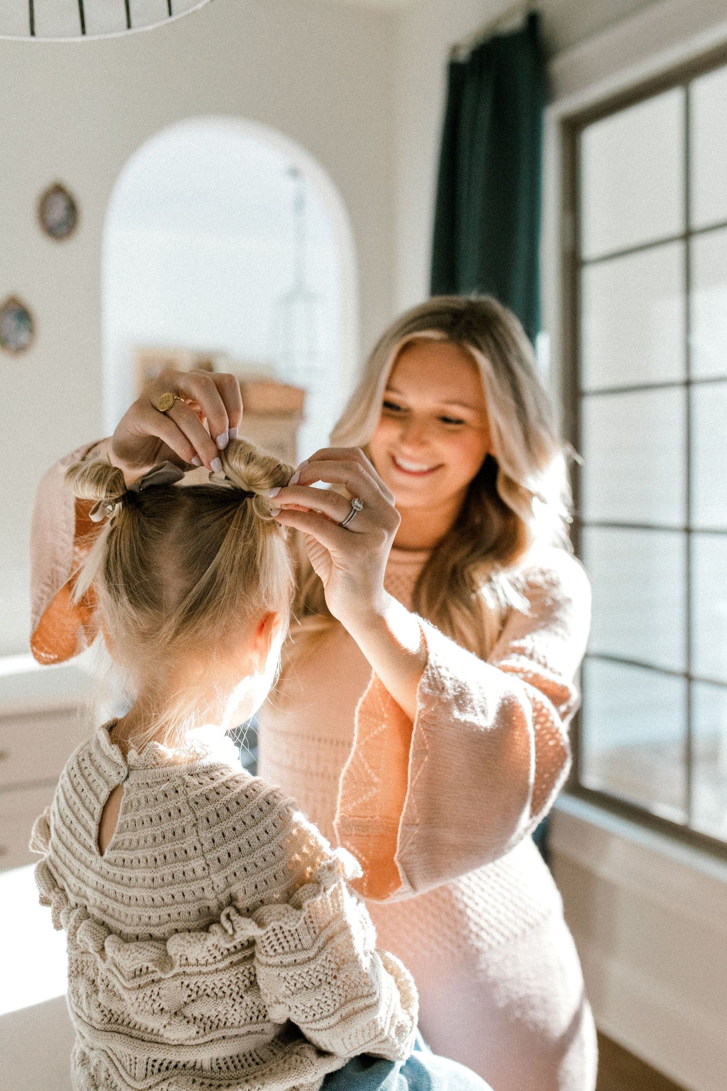 Sweet moment captured as mother styles daughter's hair in sunlit room with white walls.