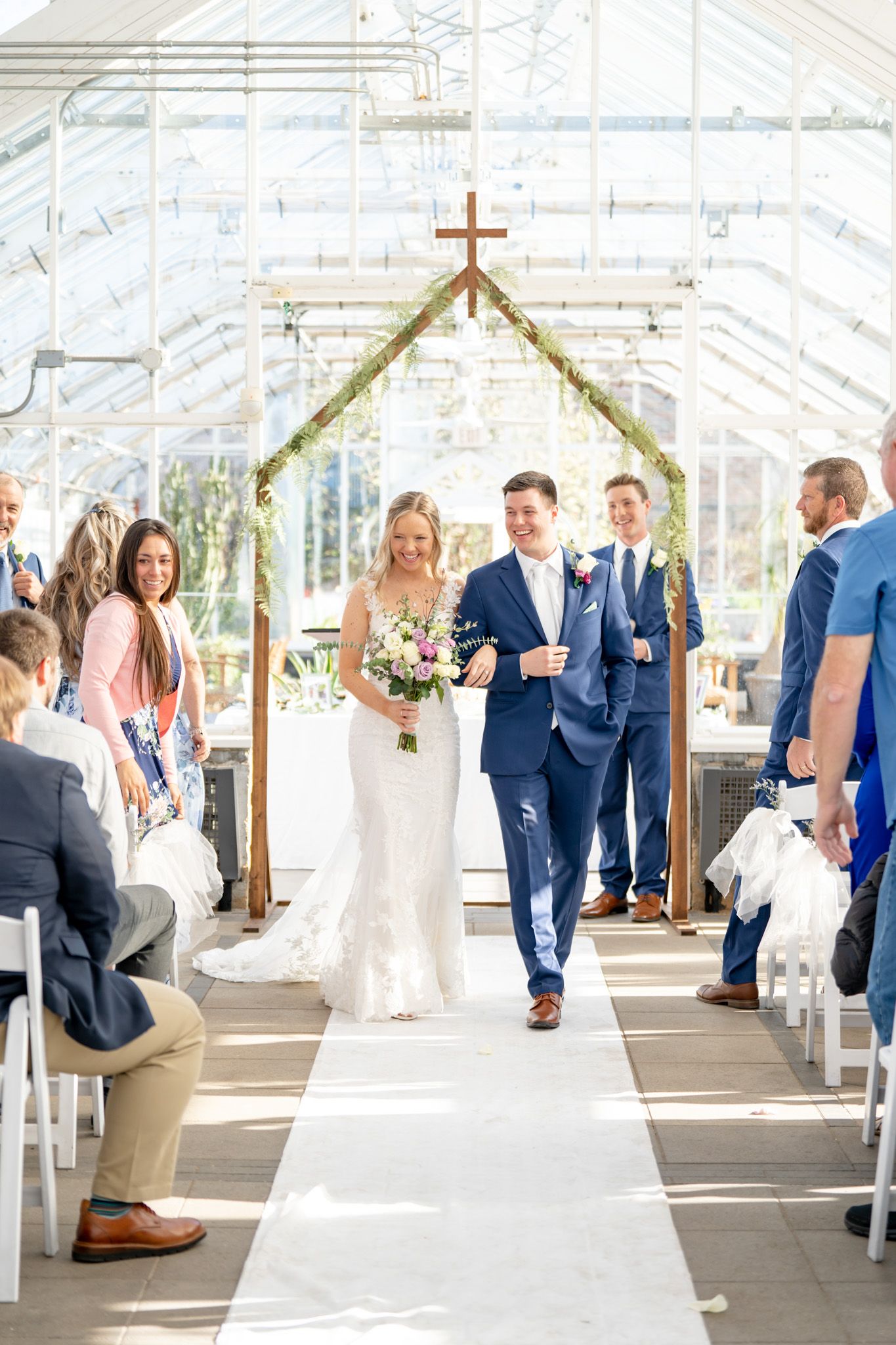Wedding ceremony aisle with white chairs and wooden cross altar under a greenhouse roof.