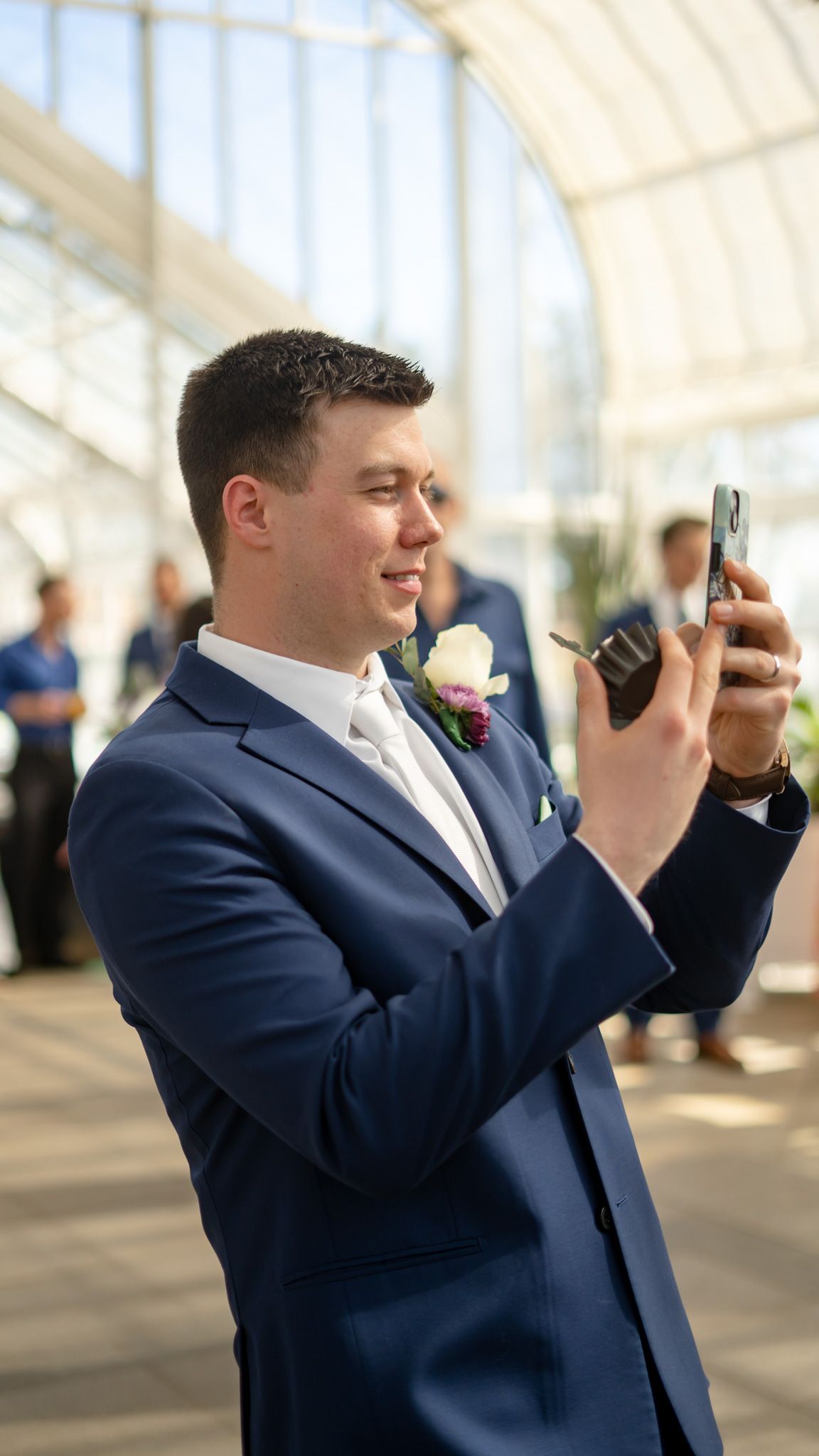 Groom in navy suit takes photos with phone while wearing boutonniere at indoor venue.