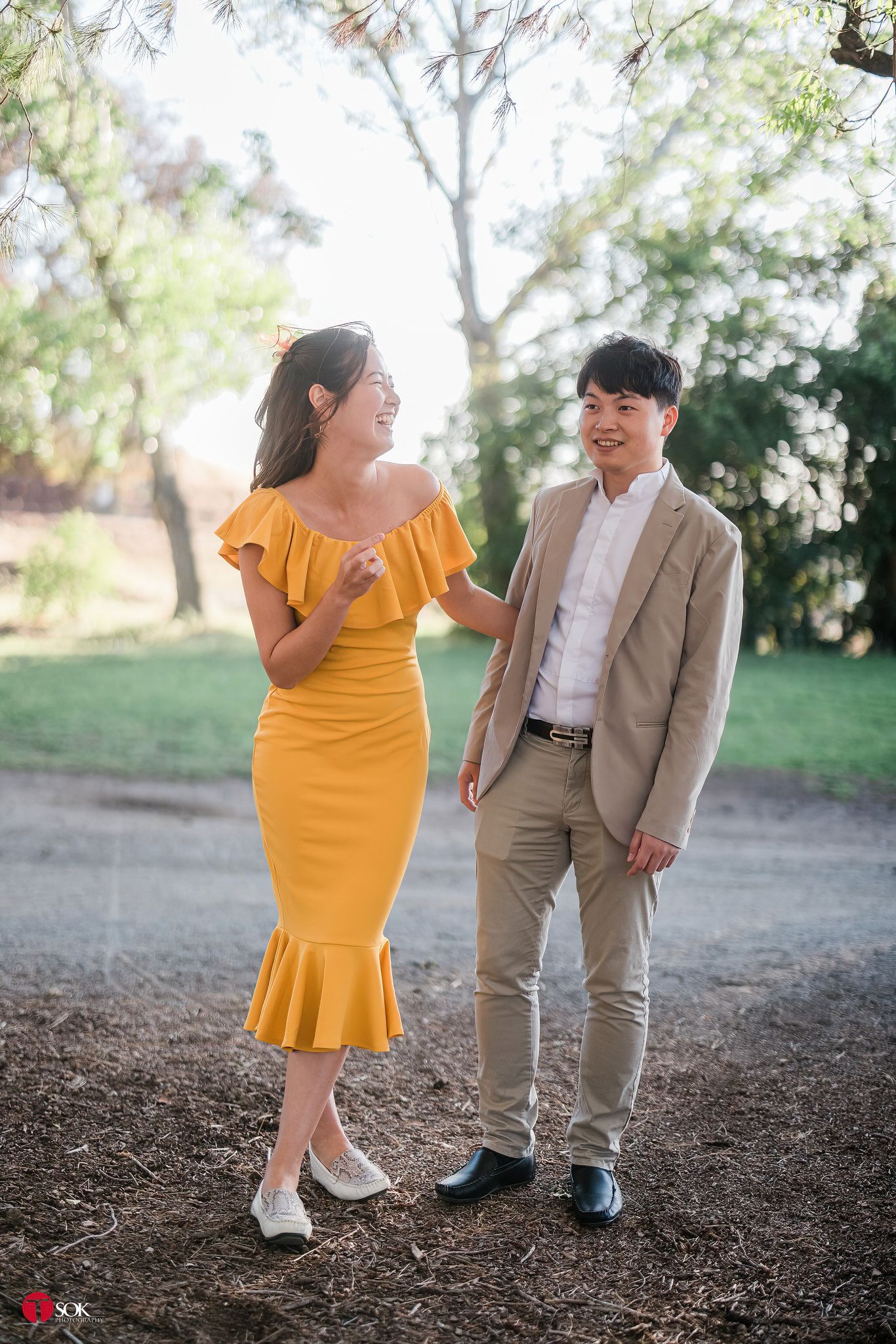 Joyful couple in yellow dress and beige suit laughing together in a park.