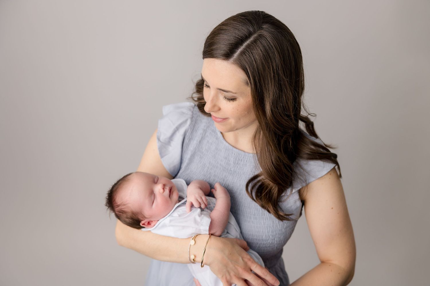A tender newborn photography session showing a mother in grey holding her sleeping baby against a neutral backdrop.