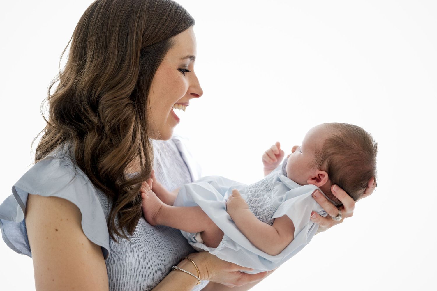 Joyful mother holding newborn baby in white outfit against bright background.