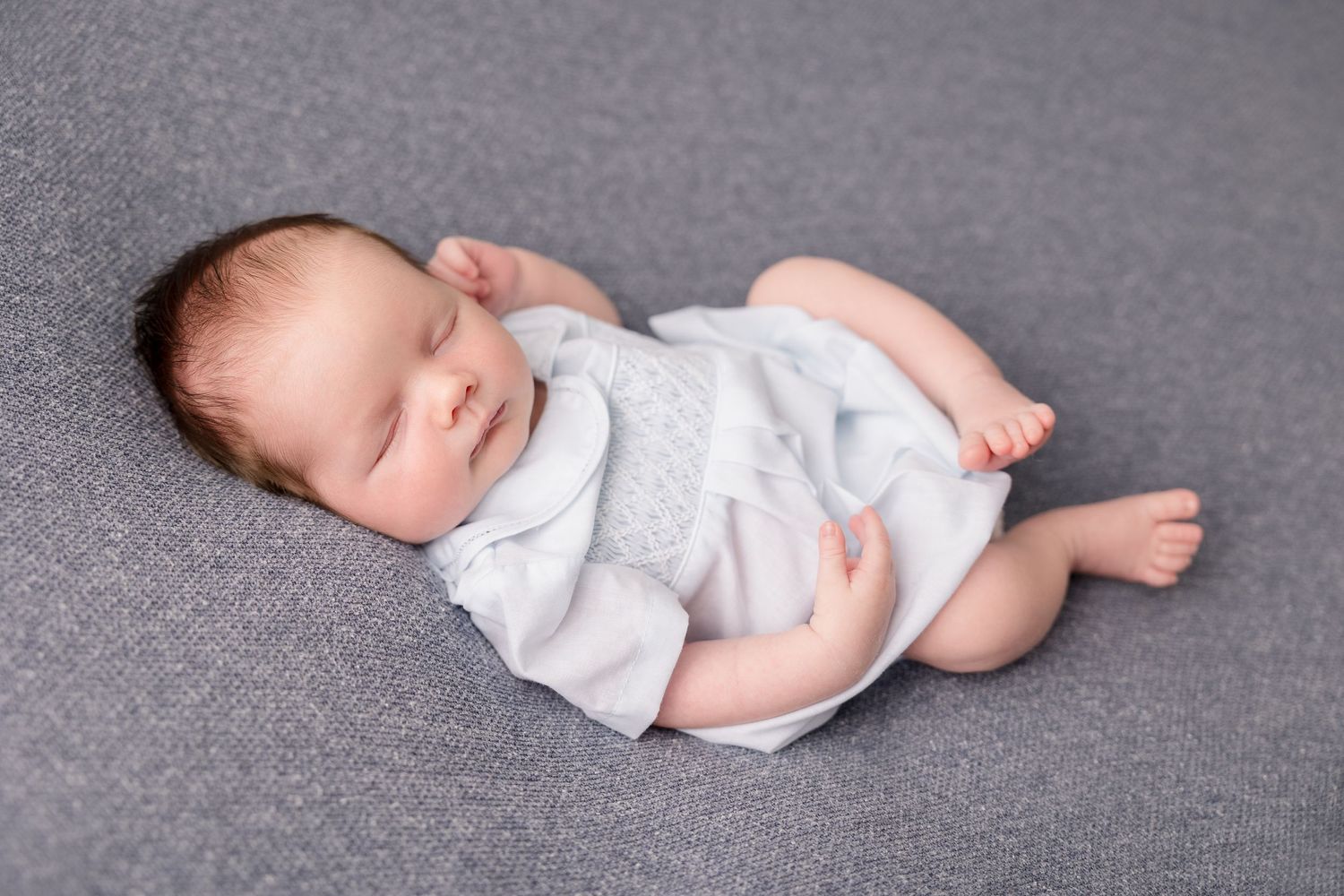 Sleeping newborn baby wearing a light grey outfit while resting on grey fabric.