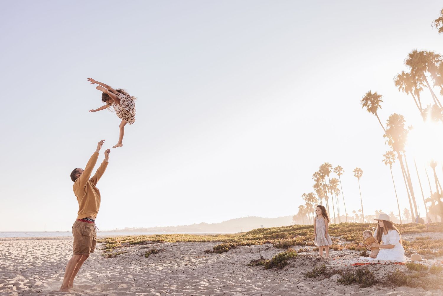 People playing and throwing child in air at sunset on beach with palm trees.