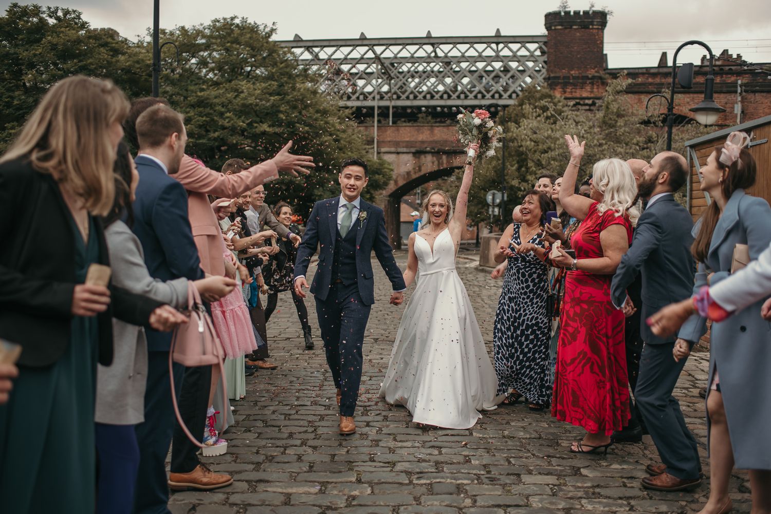Wedding celebration on cobblestone street with guests throwing confetti at the happy couple.