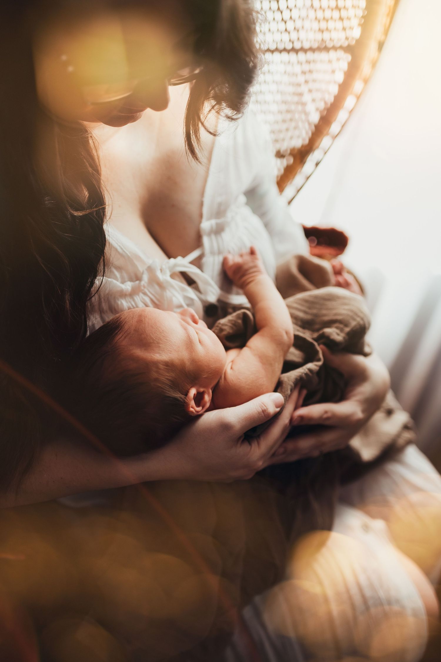 mama sits in a peacock chair by a window and prepares to nurse her newborn son at lifestyle newborn session