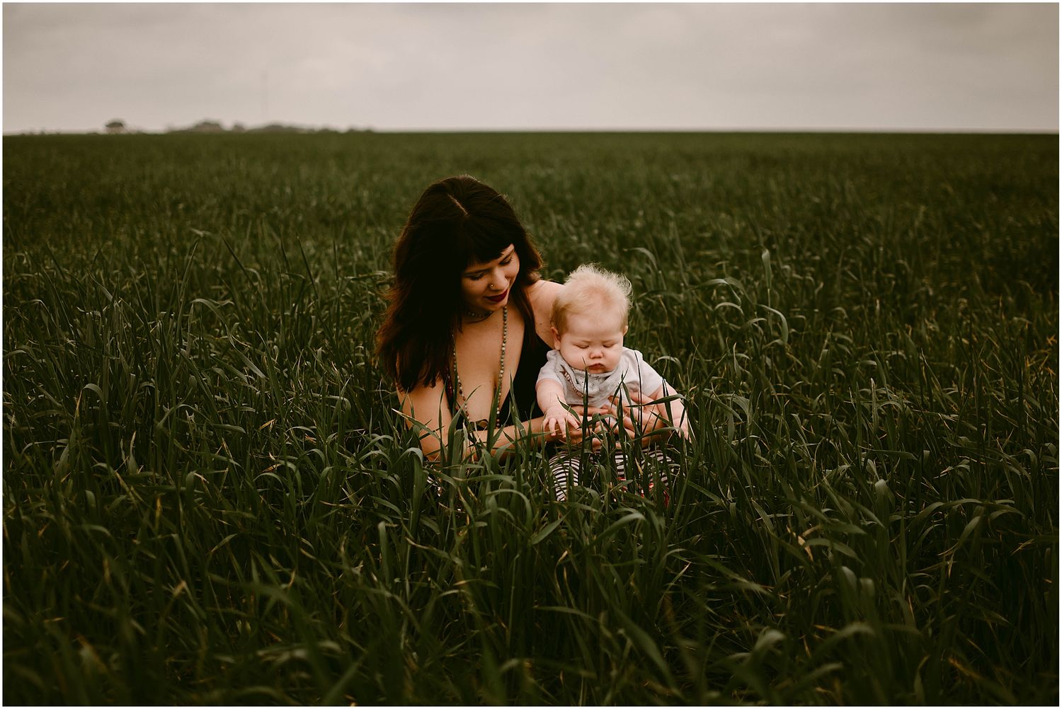 A parent and baby sit together in a lush green field under an overcast sky.