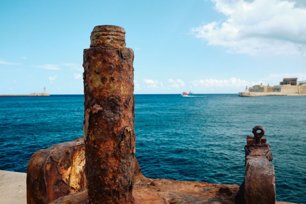 Rusty iron bollards on harbor edge with blue Mediterranean waters and fortress in background.