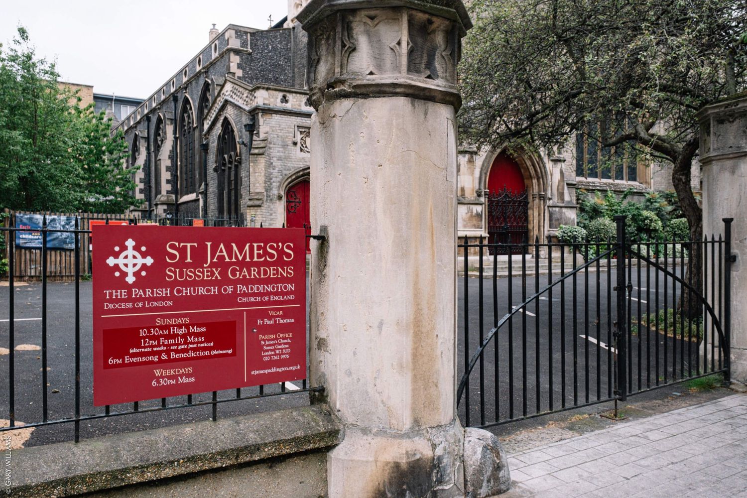 Red sign and entrance gate for St James Church with stone columns and iron fencing.