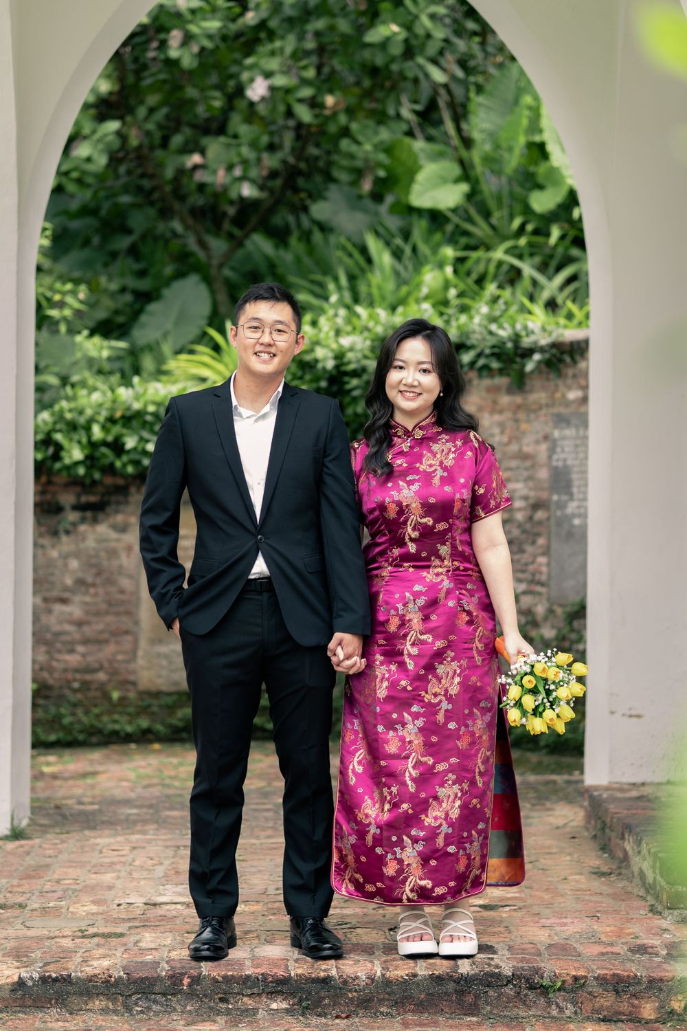 Couple standing under white archway in garden setting wearing formal attire.