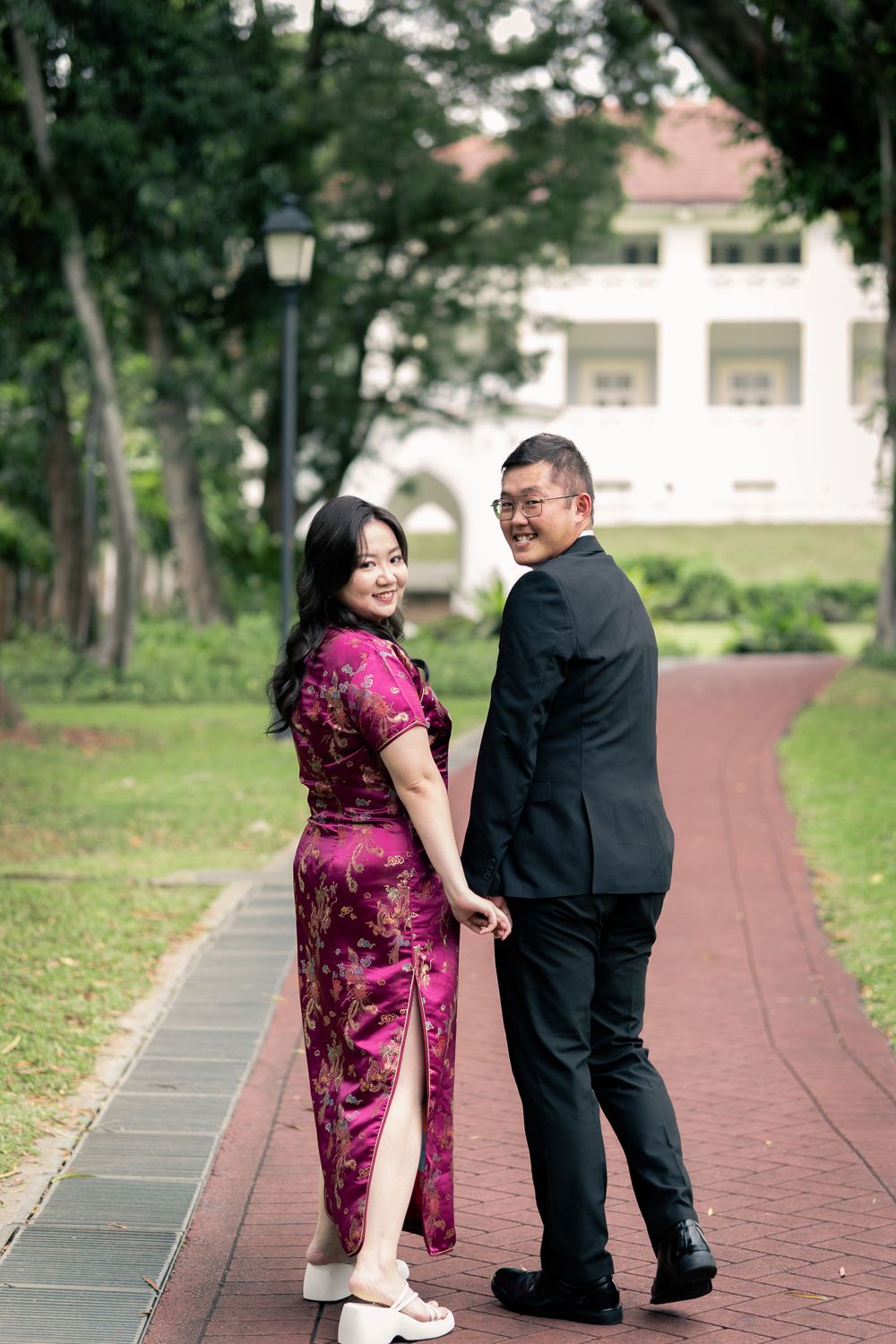 Couple in formal attire looking back at camera while walking on a red brick path.