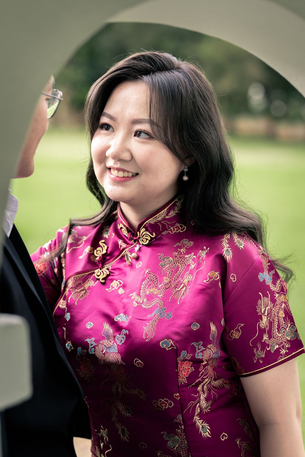 A person wearing a traditional pink and gold floral qipao dress smiles warmly outdoors.