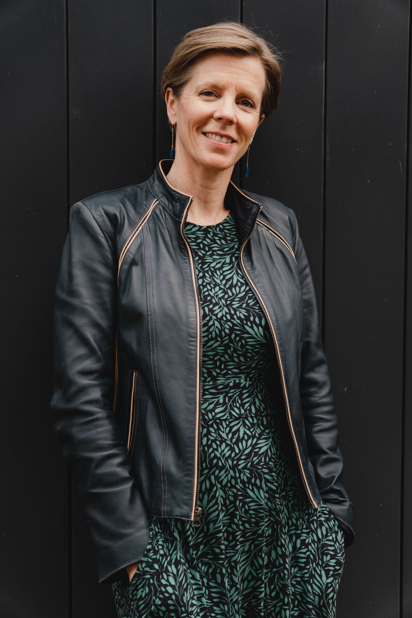coach woman standing against black wall smiling wearing black jacket and dress