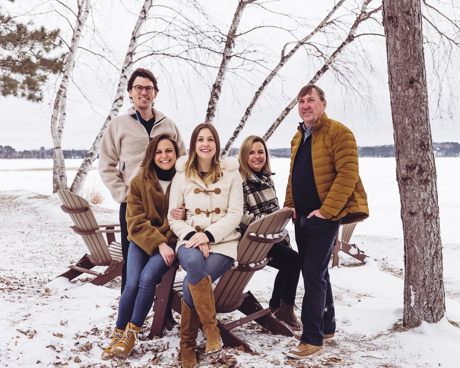 Family group in winter attire poses together on snowy beach with birch trees.