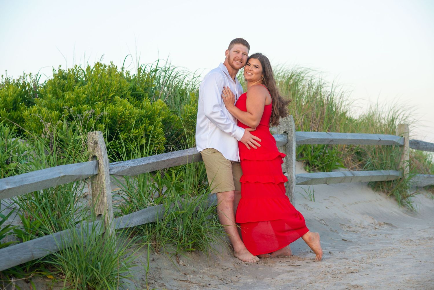 A couple embraces by a wooden fence on a beach path with green foliage at sunset.