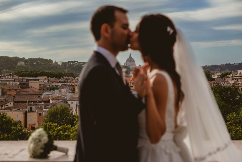A wedding couple embraces with a scenic Italian cityscape visible in the background.