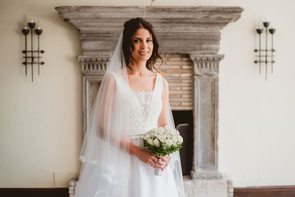 A bride in a flowing white gown stands elegantly in front of a rustic stone wall with architectural details.
