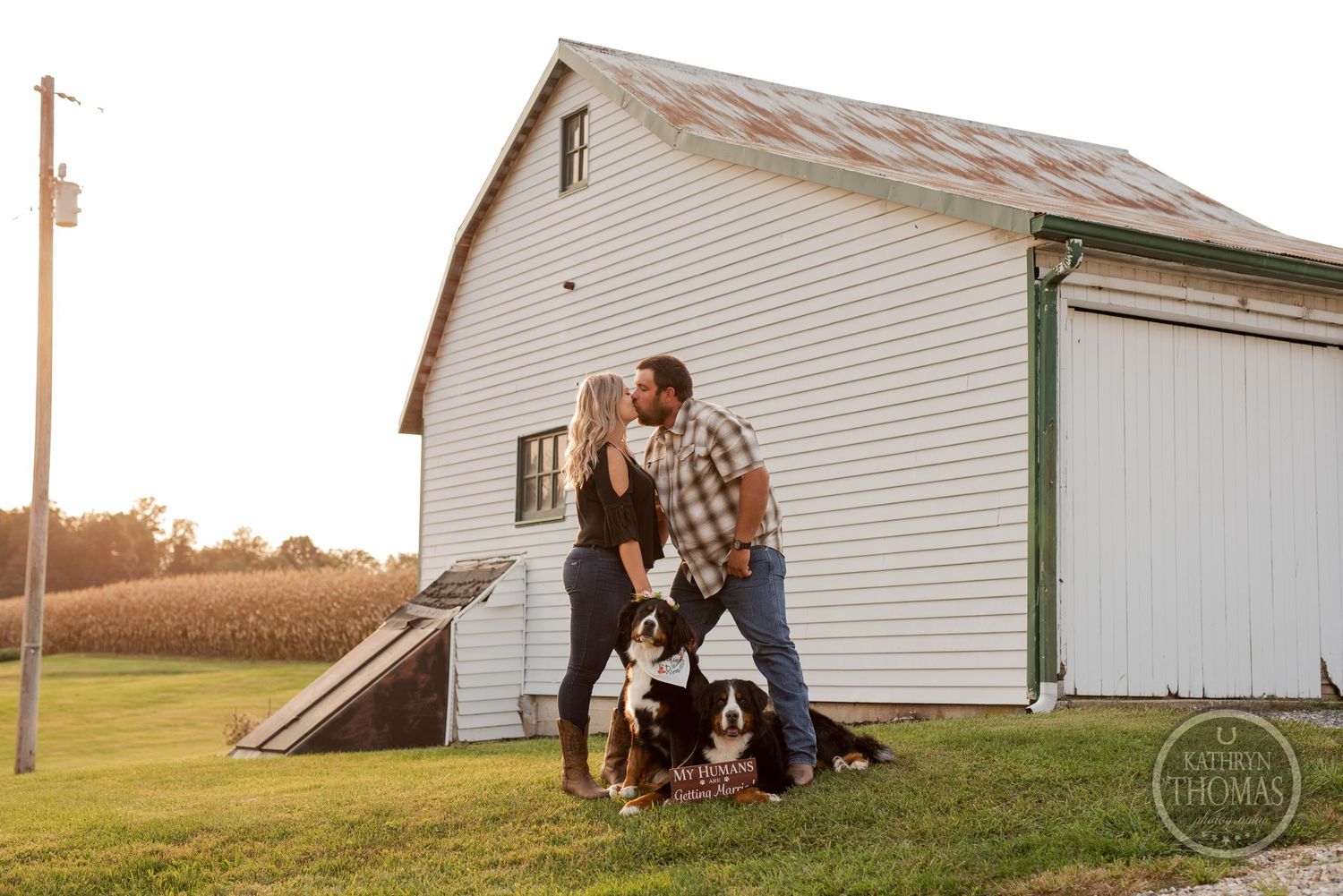 A couple and their dogs share a tender moment outside a white barn at sunset.