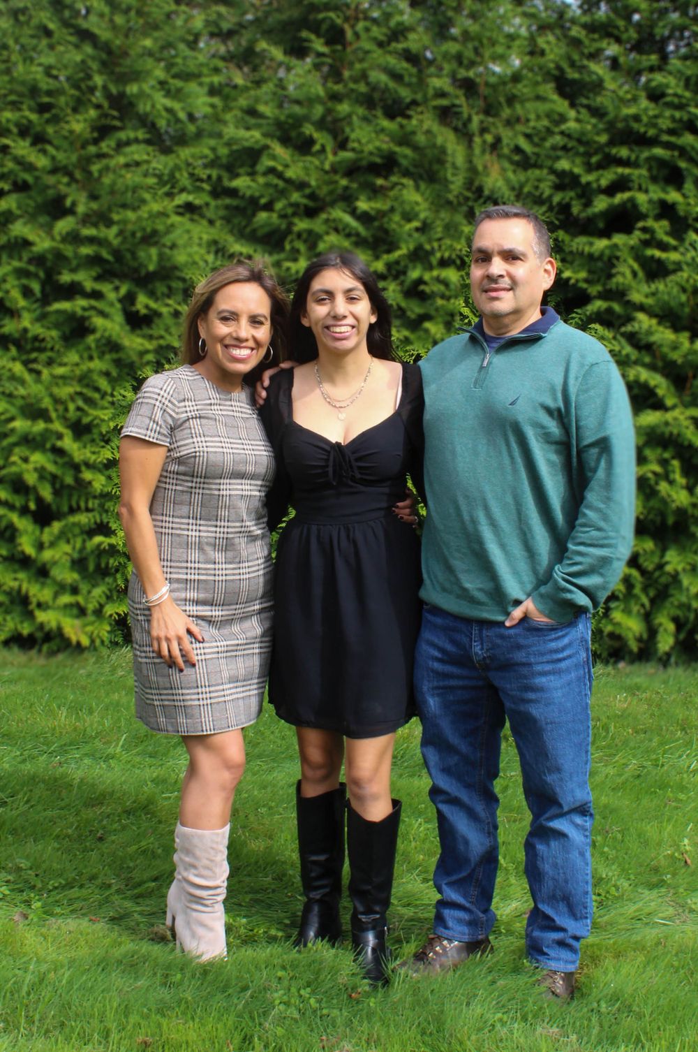 Three people pose together on grass with green hedge backdrop.