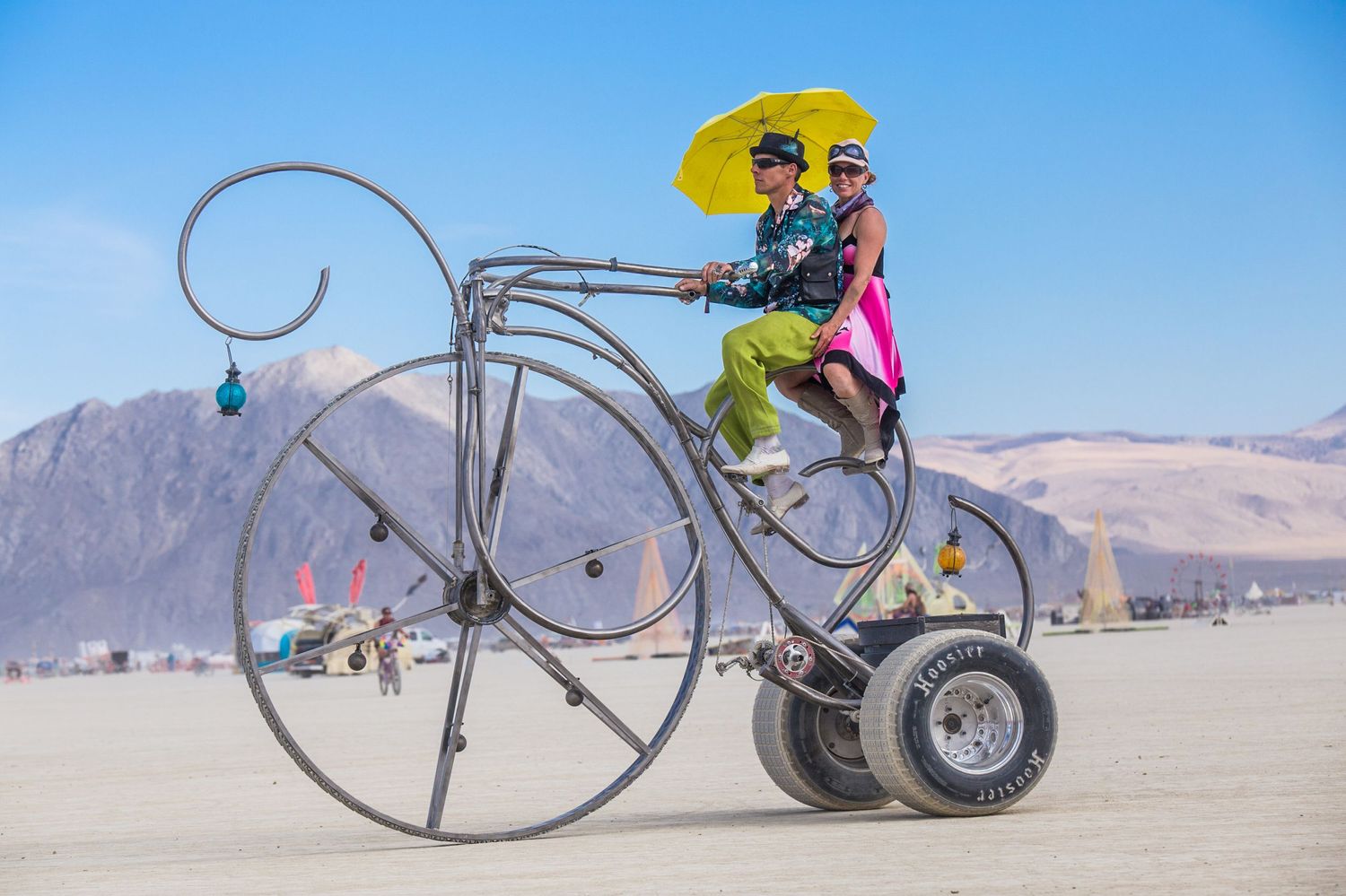A whimsical art bicycle sculpture with oversized wheels and unique design stands in a desert landscape with mountains at Burning Man