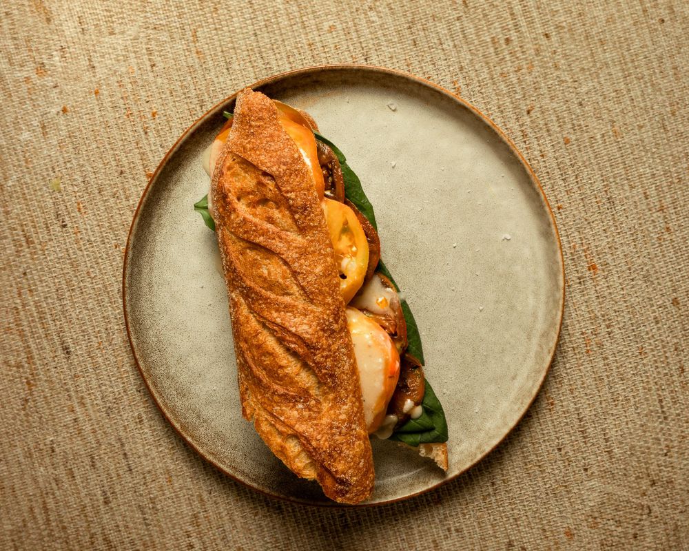 Gourmet sandwich on crusty baguette with meat and vegetables served on beige ceramic plate.