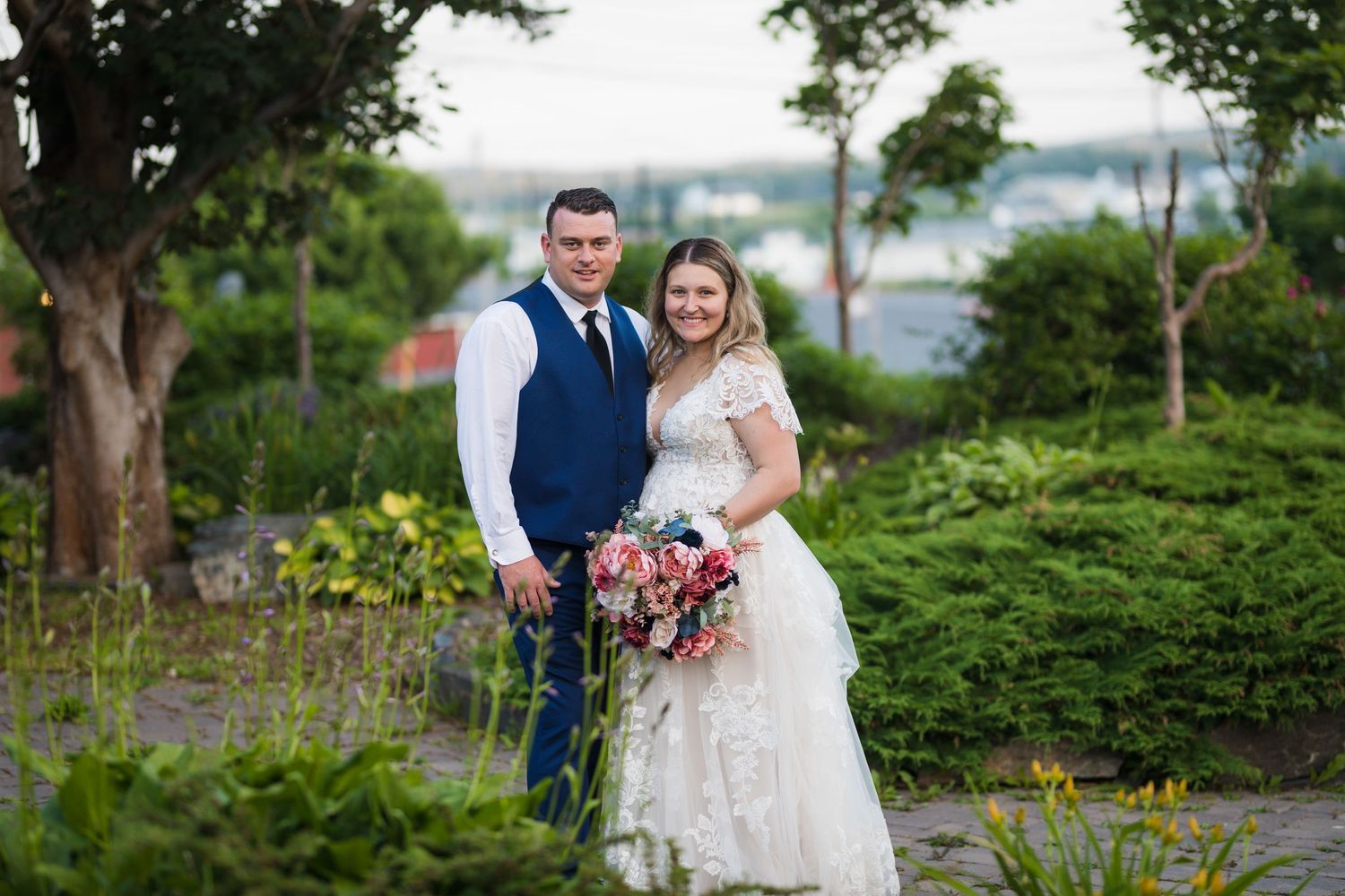 Wedding couple poses in garden setting with colorful bouquet and lace dress details.