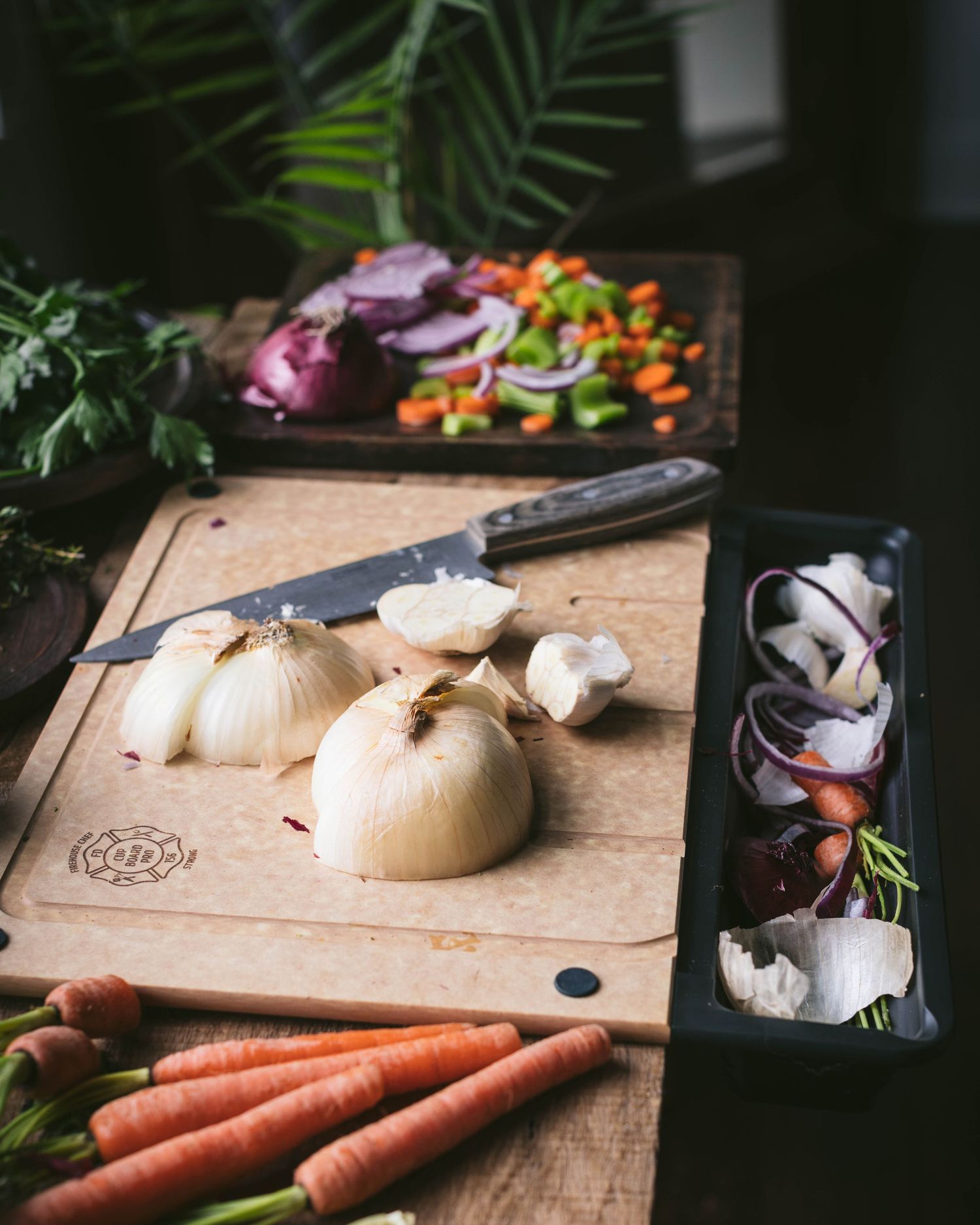 Onions and mushrooms on a wooden cutting board with carrots and colorful diced vegetables in the background.