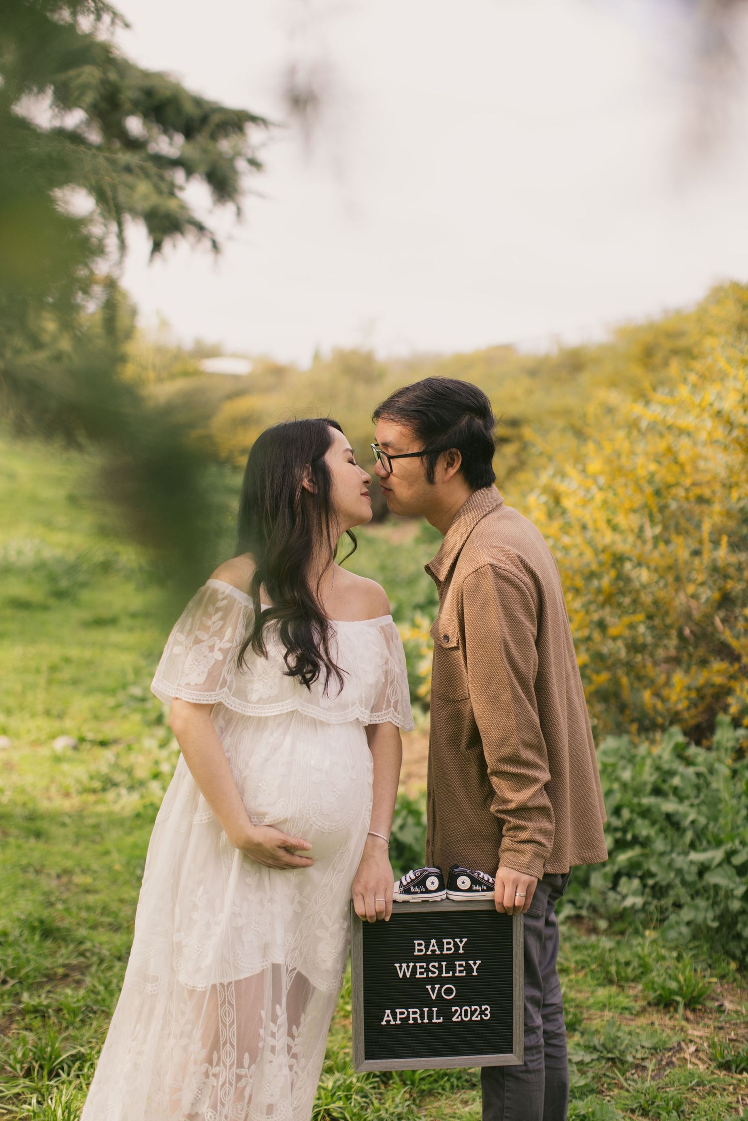 Couple shares intimate moment during pregnancy announcement with letterboard in forest setting.