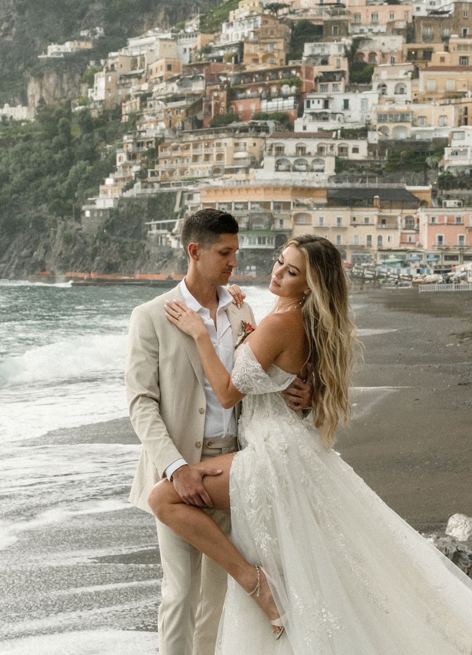 Romantic wedding photo on Positano beach with coastal buildings in background.
