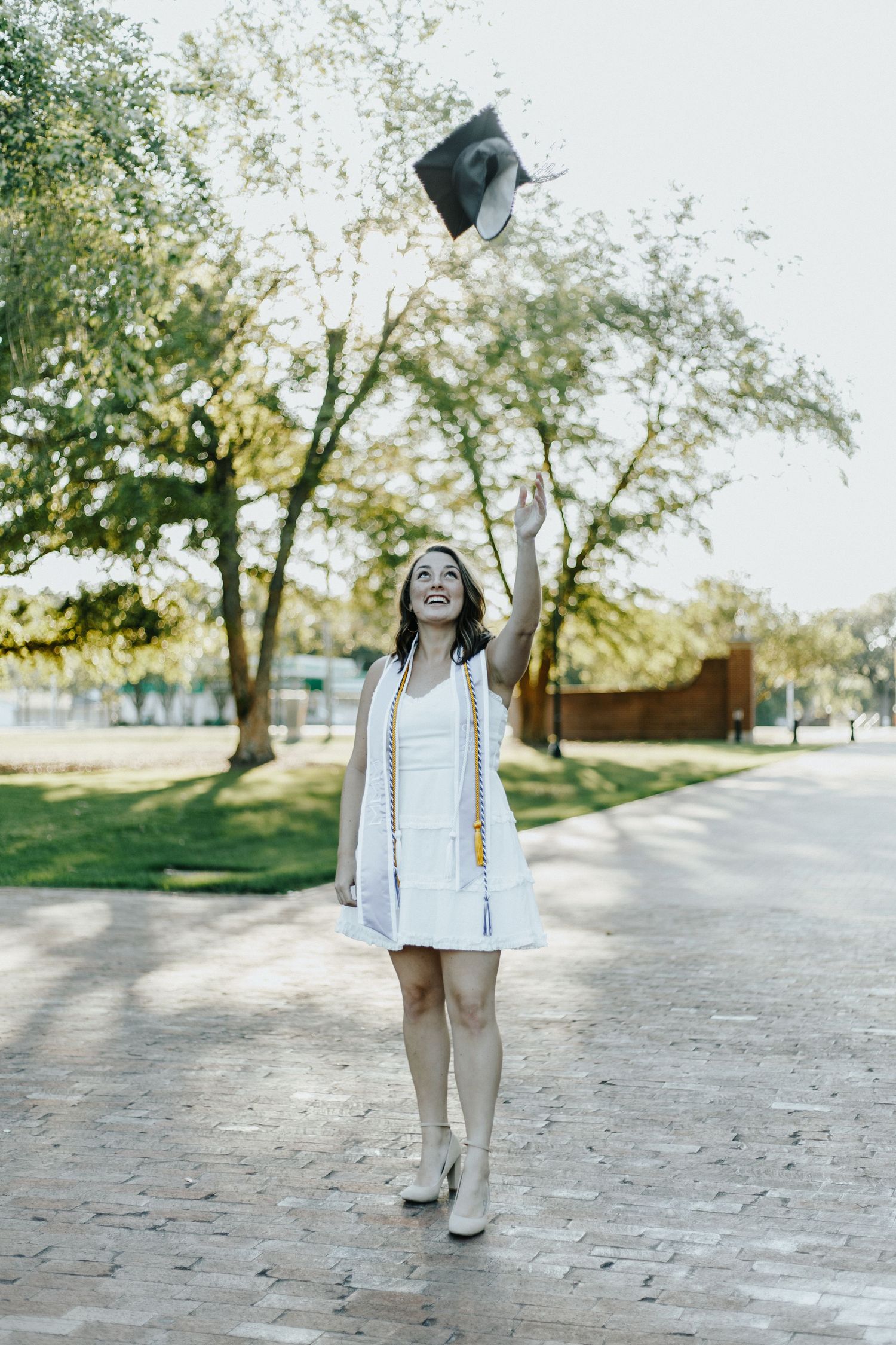 Graduate in white dress tossing cap in air during outdoor celebration.