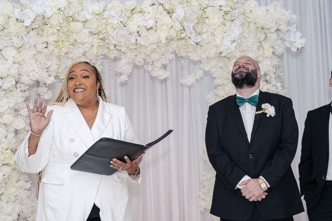 Wedding ceremony in progress with white floral wall backdrop and officiant in white suit.